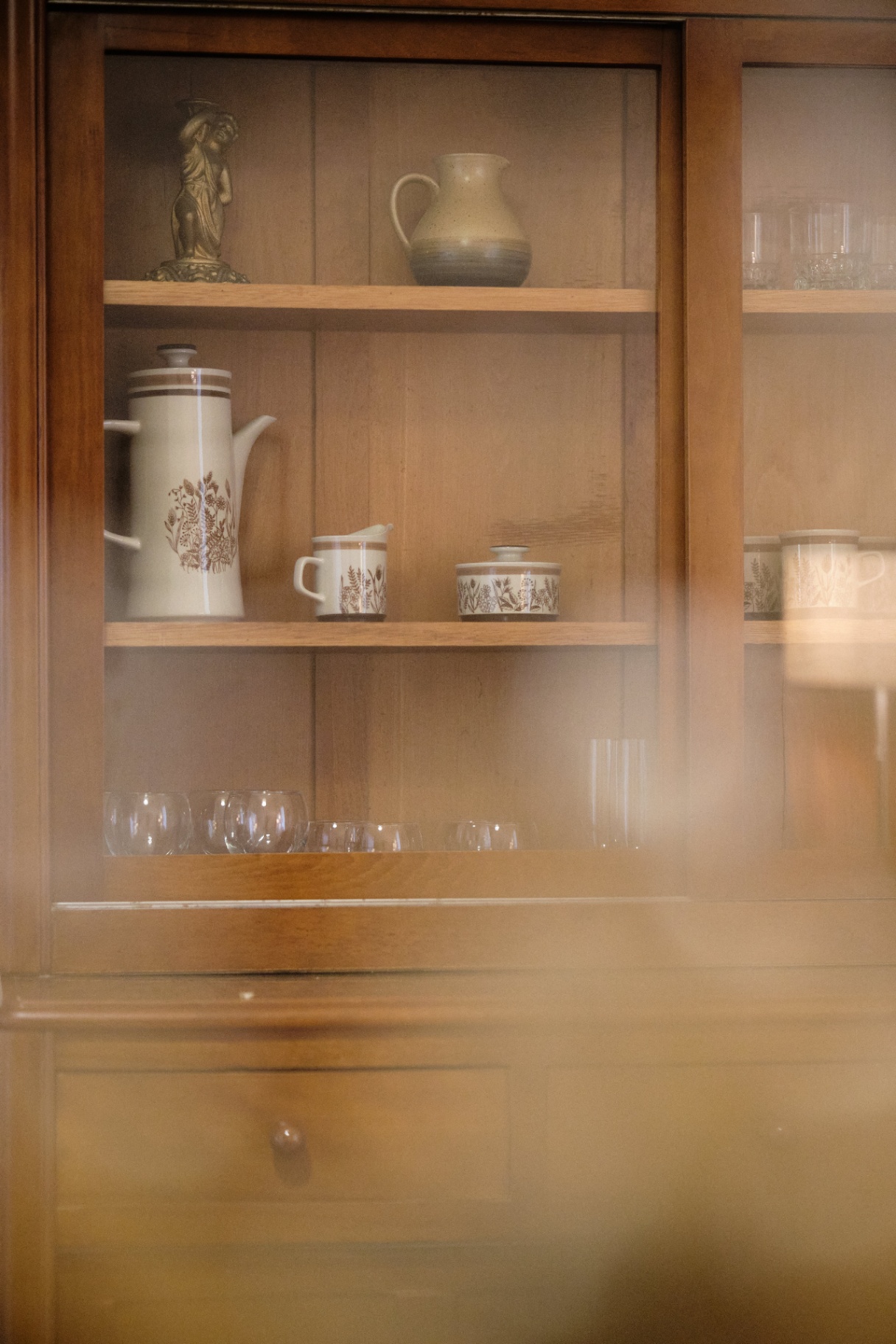 Glass-fronted wooden cabinet displaying heirloom stoneware coffee set, pottery jug, and crystal glasses.