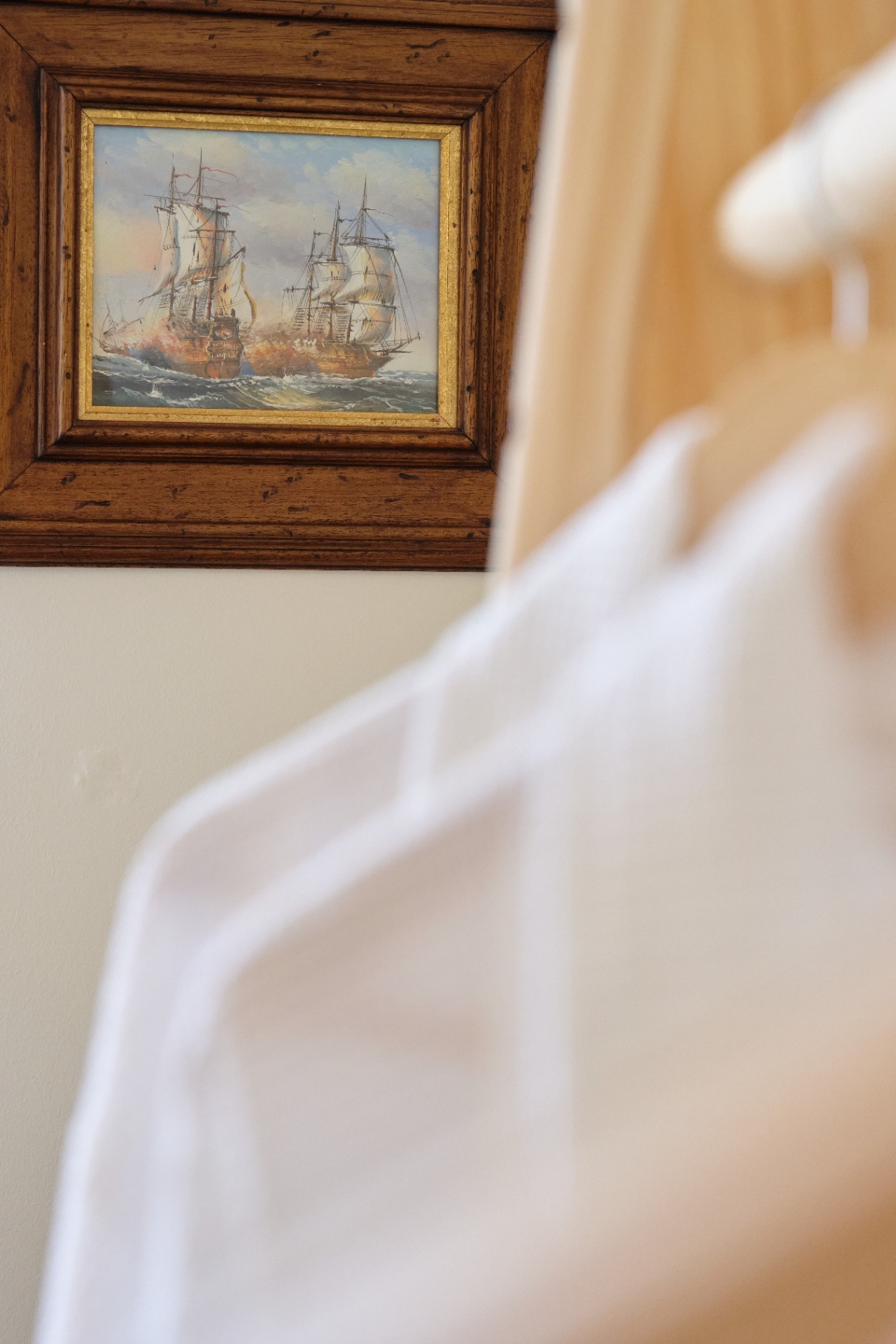 Framed oil painting of tall ships at sea hung on a soft cream wall above crisp white linens.