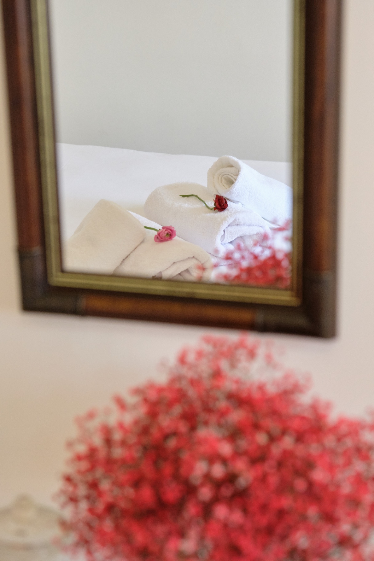 Rolled white towels with a rose reflected in a wooden-framed mirror above blush gypsophila.