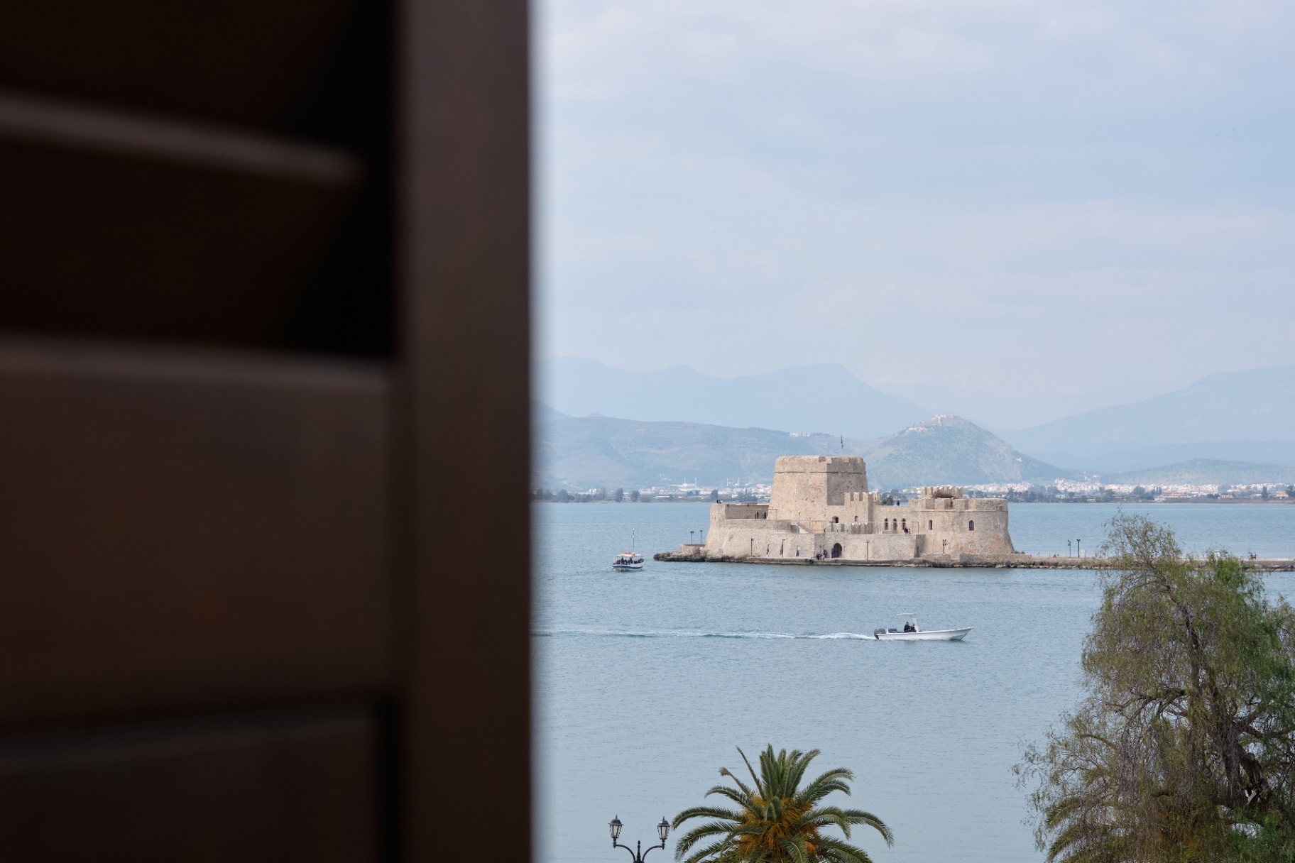 View of the Bourtzi fortress in Nafplio's bay framed through a shuttered window.
