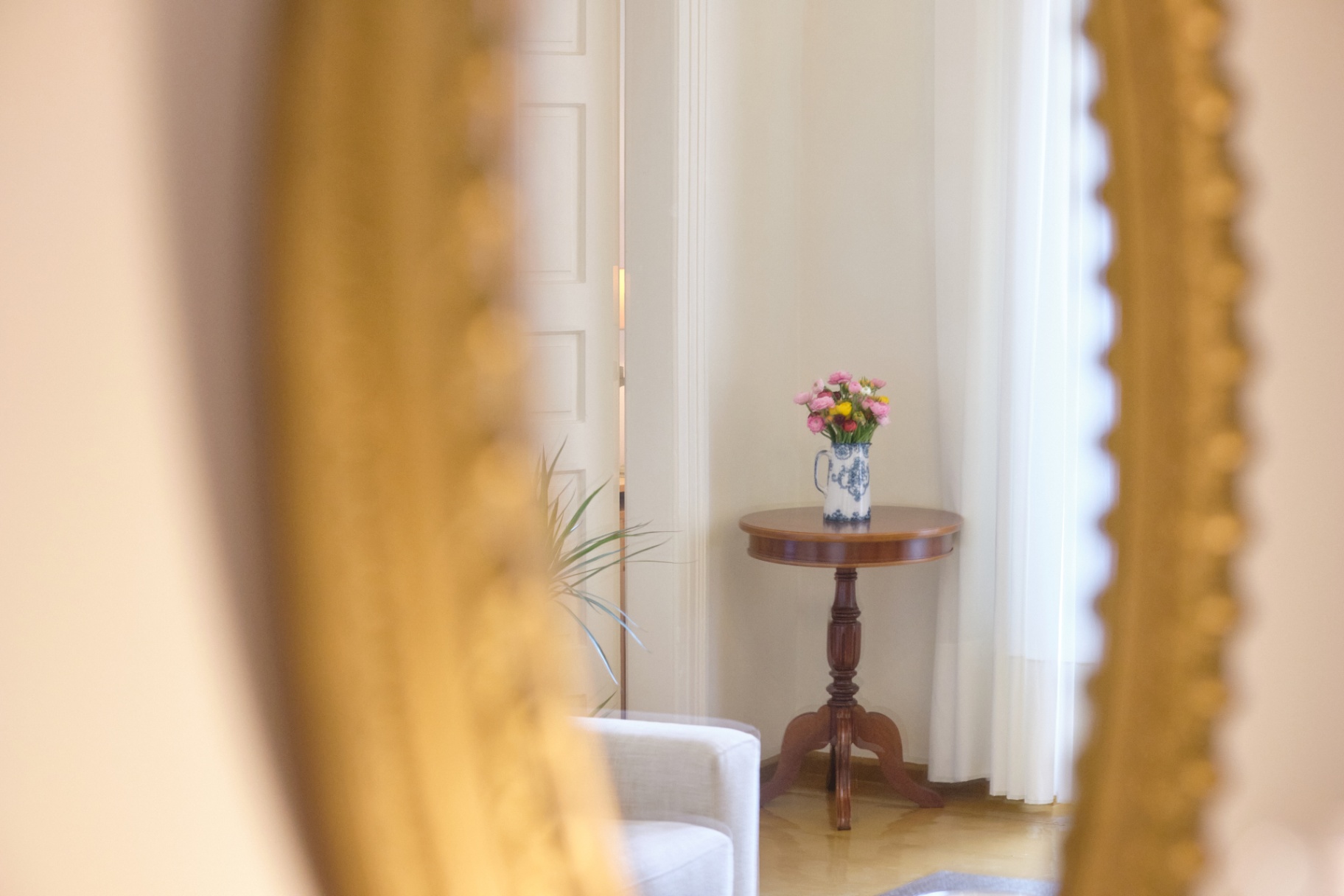 Reflection in a gilt mirror revealing a pedestal table with a blue-and-white pitcher of flowers.