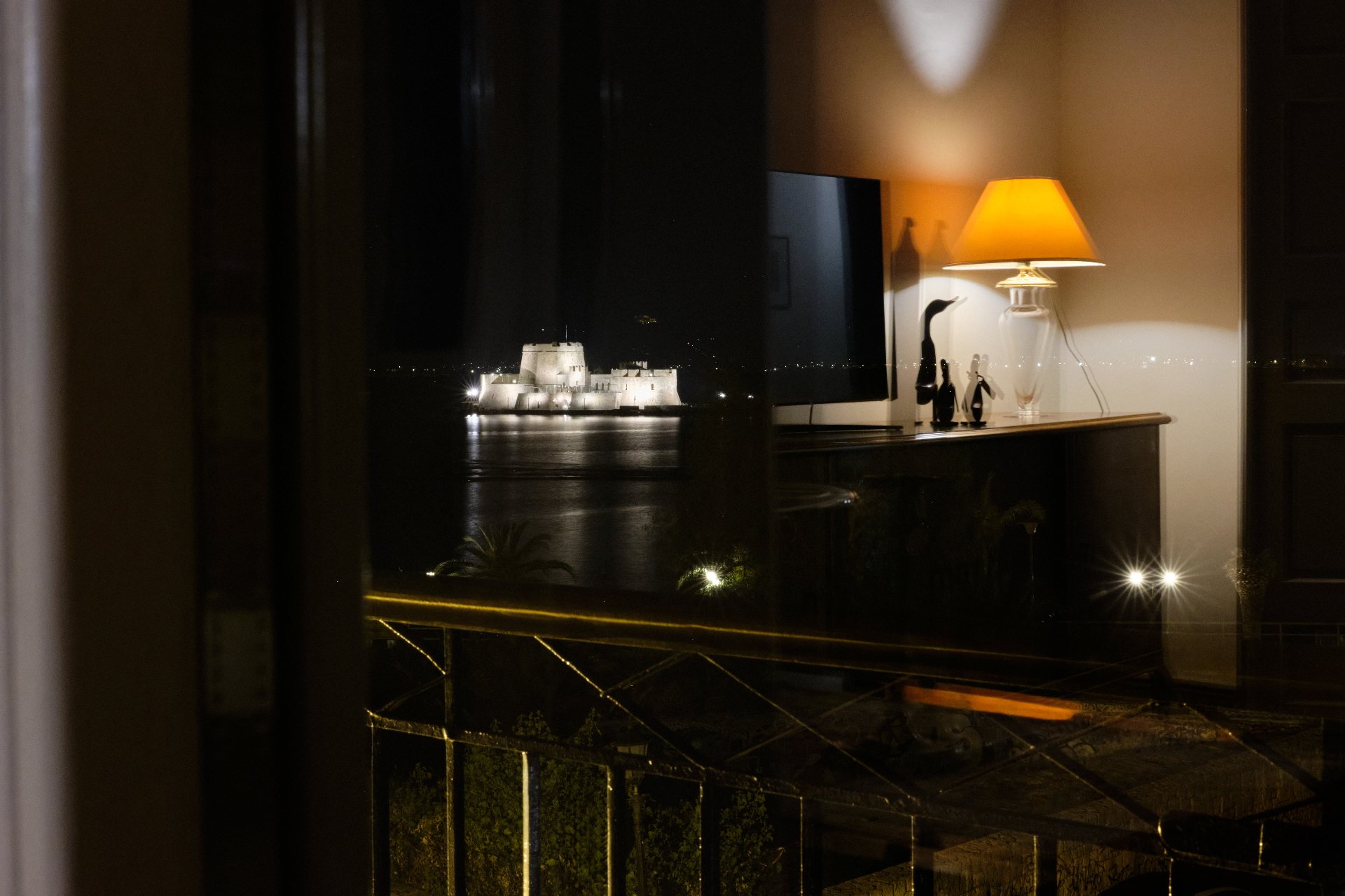Glowing bedside lamp framing a window view of the floodlit Bourtzi fortress across the dark bay at night.
