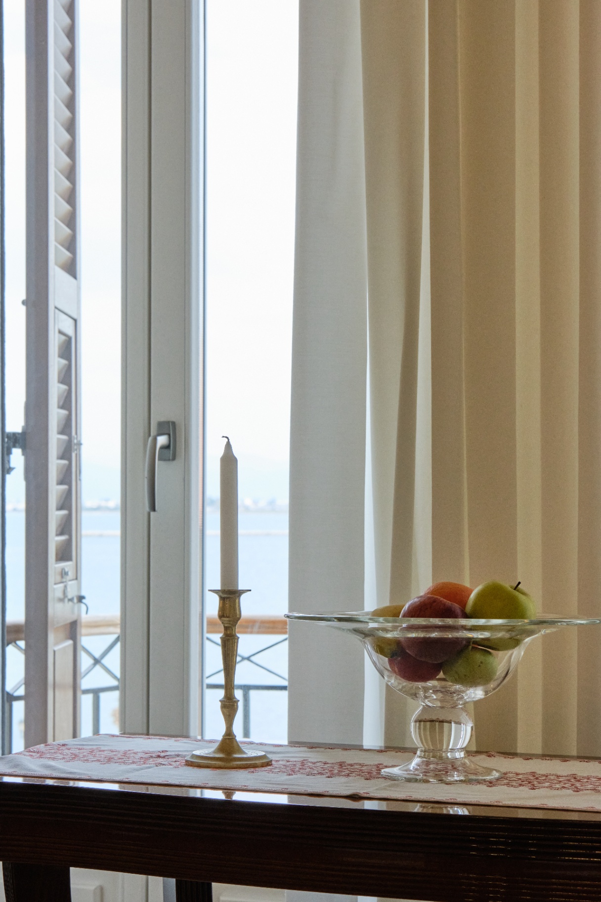 Brass candlestick and glass fruit compote on an embroidered runner before a sea-view balcony.
