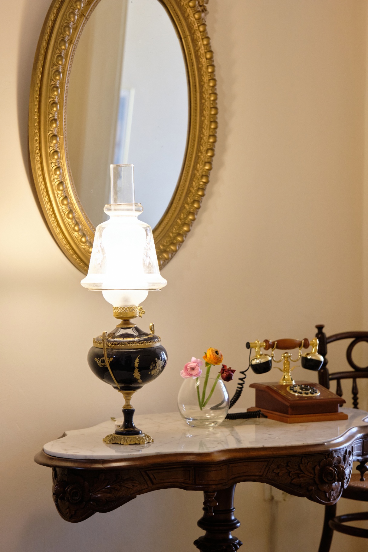 Cobalt-and-gold porcelain oil lamp and antique brass telephone on a marble-top table beneath a gilt oval mirror.