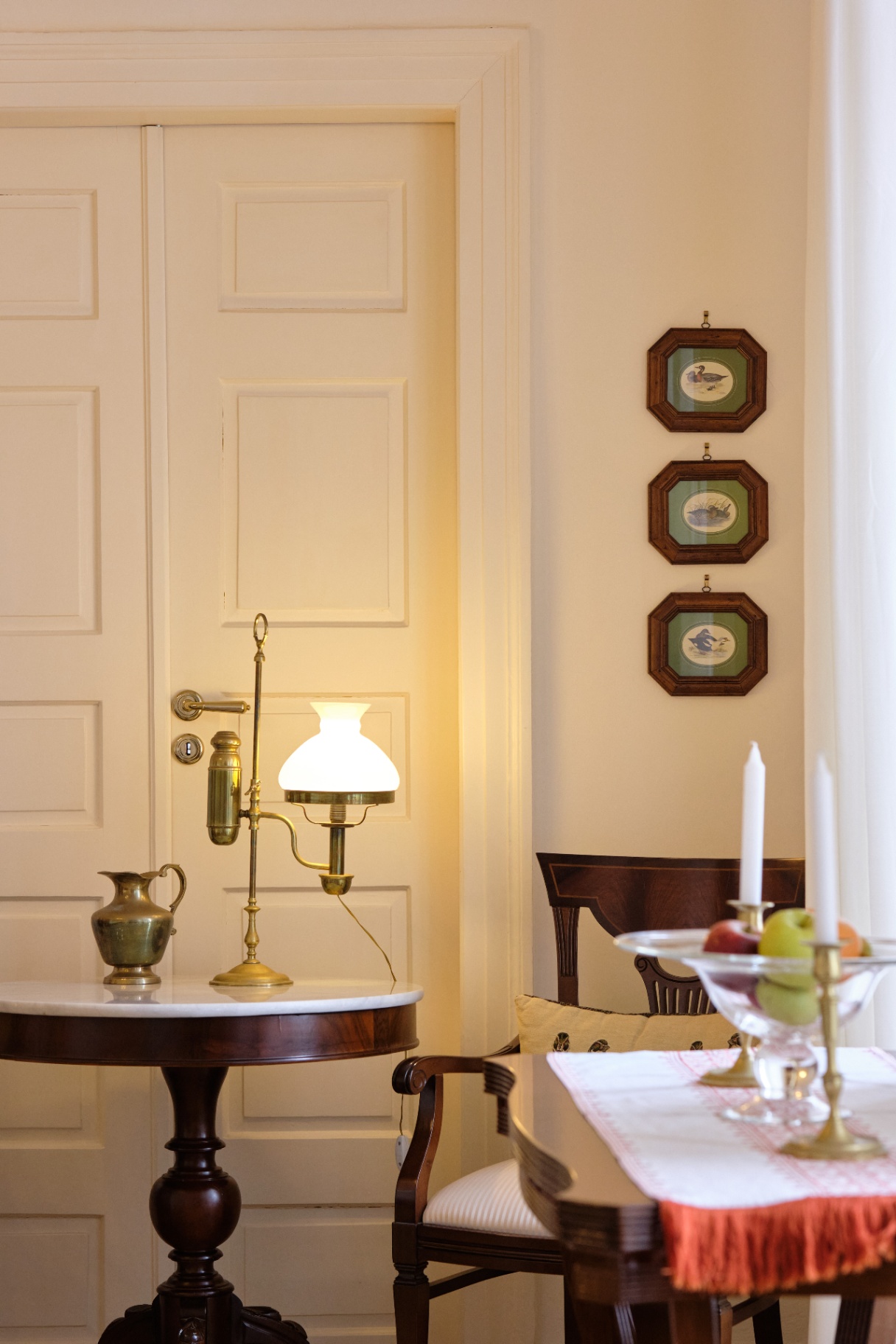 Antique brass reading lamp on a marble-top table beside a paneled door and framed bird prints.