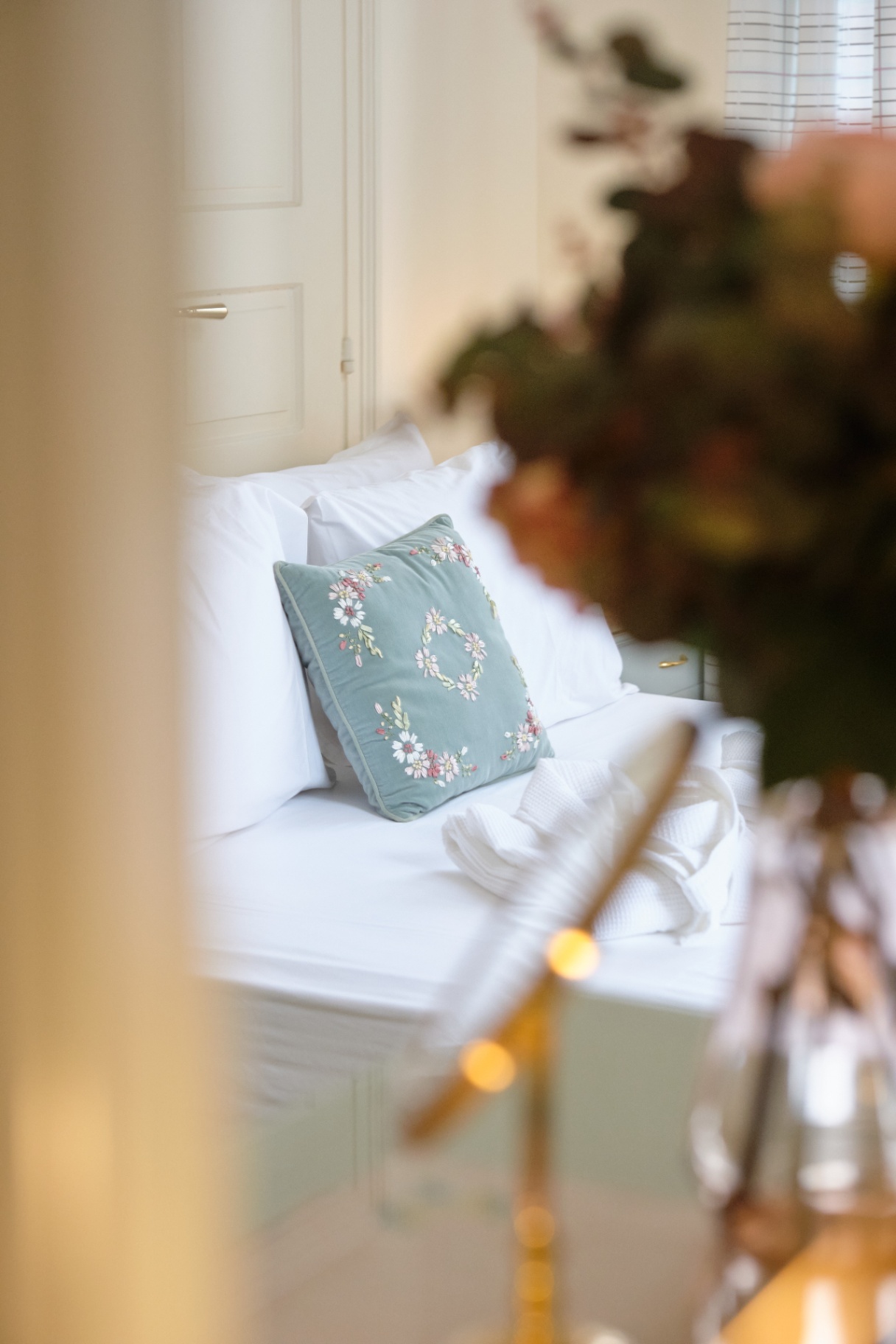 Soft bedroom vignette with white linens and a pale blue embroidered cushion seen past a bouquet.