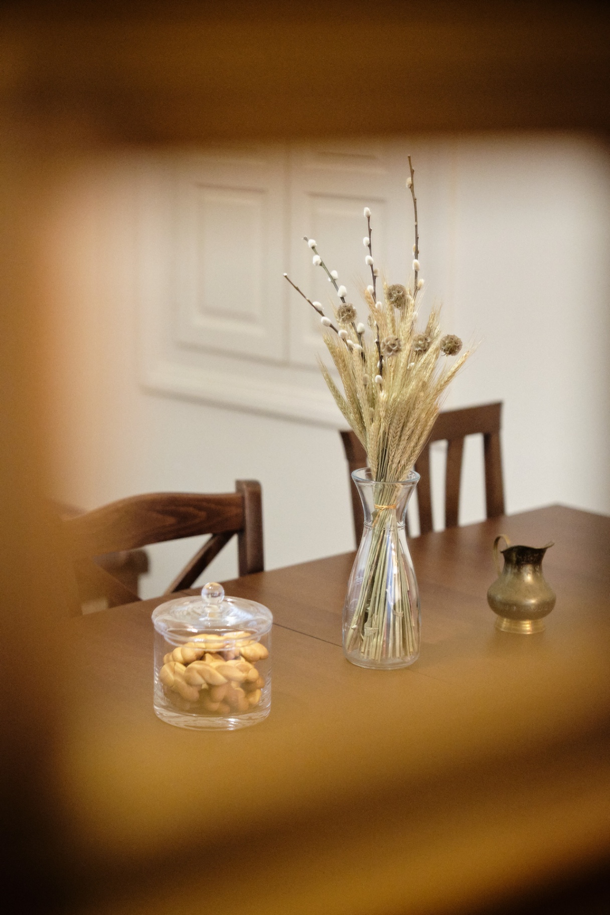 Dining table set with a glass jar of koulourakia, wheat-and-pussy-willow bouquet, and antique brass jug.