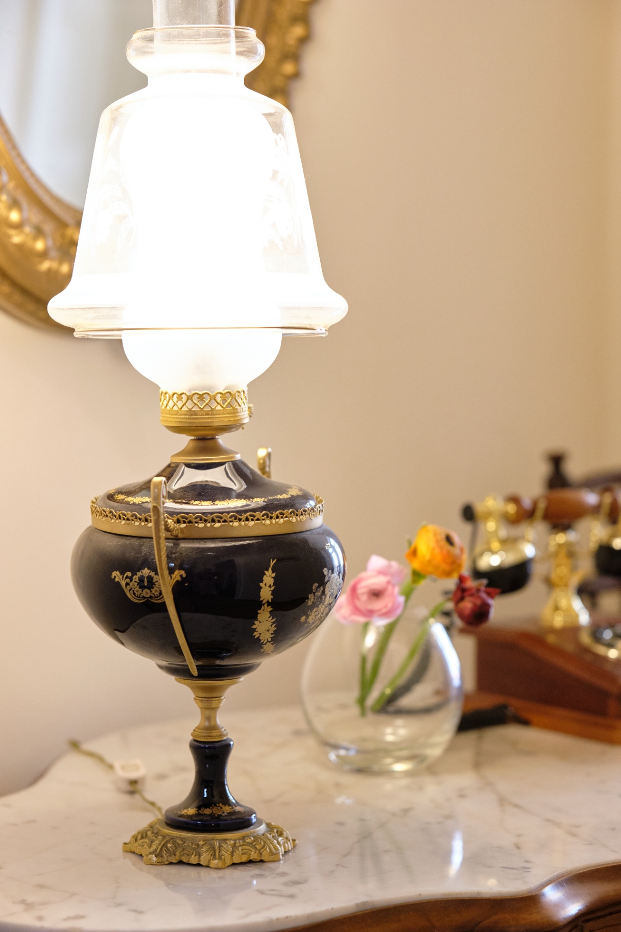 Cobalt-and-gold oil lamp with frosted glass shade on a marble table beside fresh flowers.